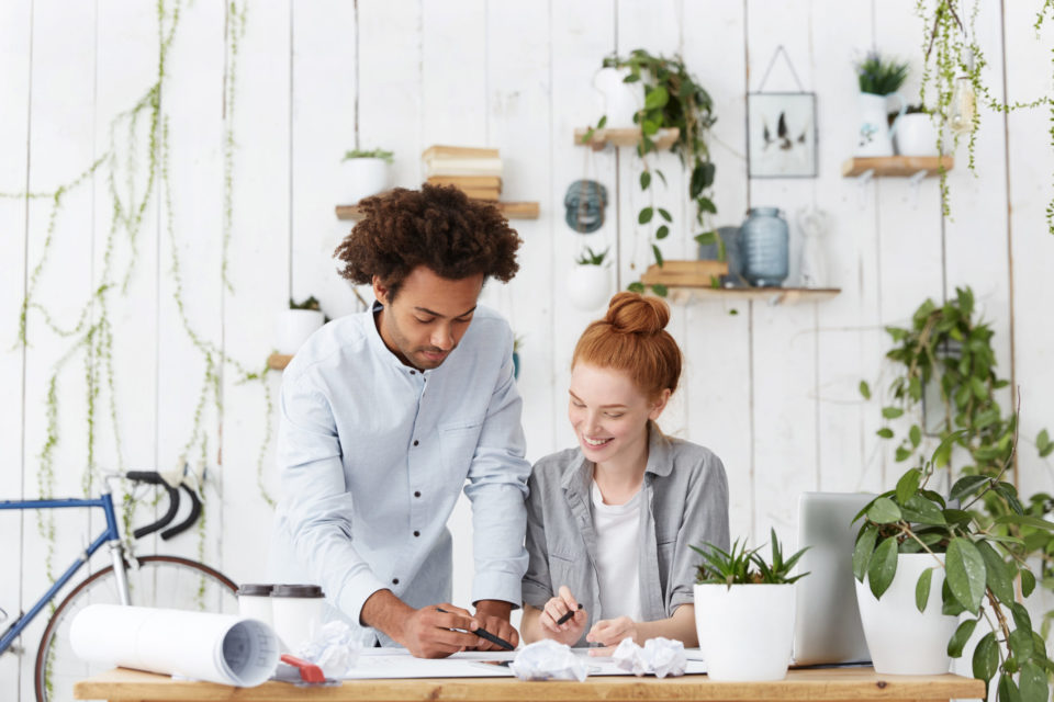 Teamwork and cooperation concept. International team of two young ambitious designers working on common project together, smiling, brainstorming, enjoying creative process in cozy office interior